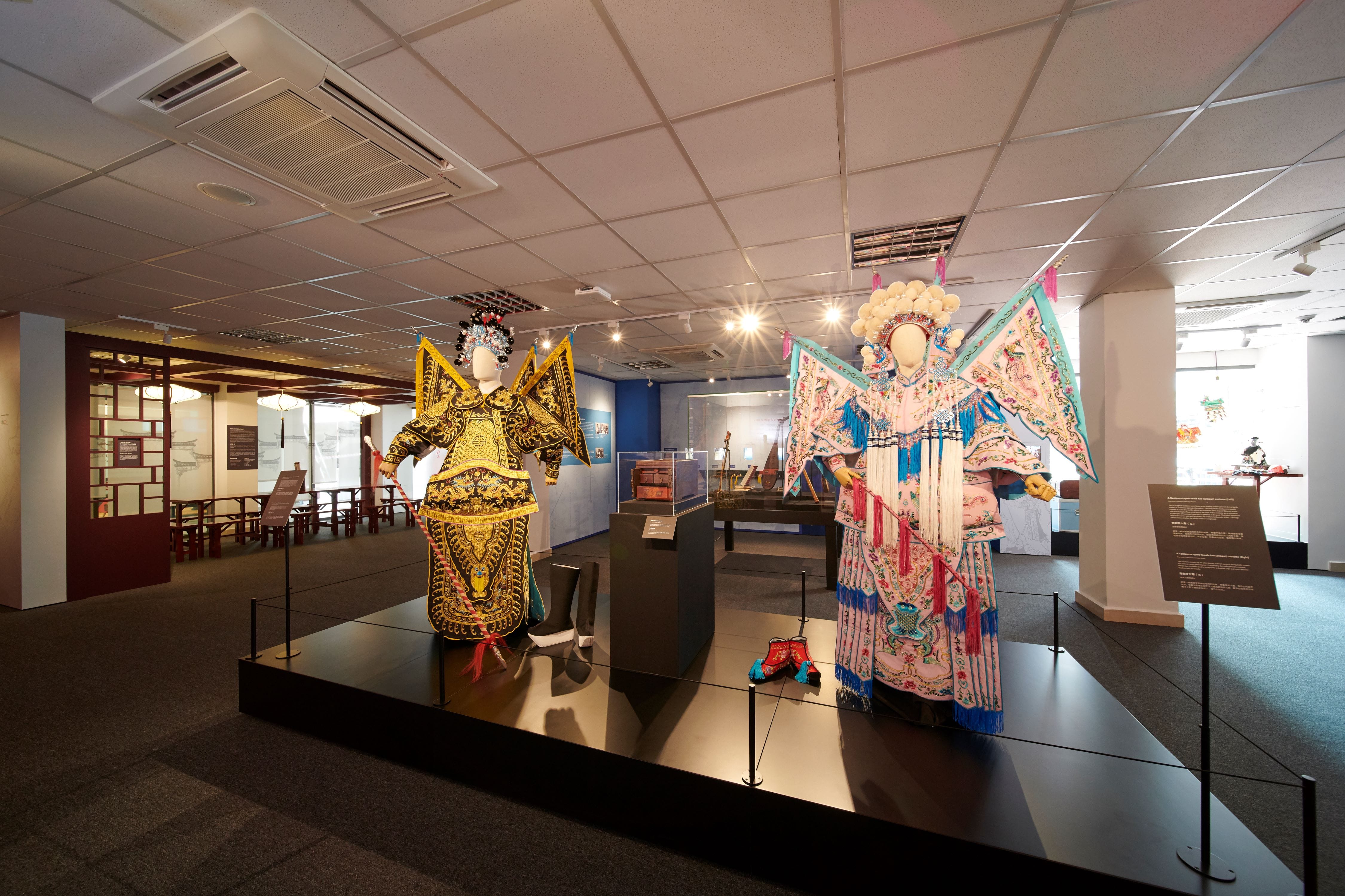 Two mannequins in elaborate Chinese opera costumes, one gold and one pink with teal, displayed on a raised platform in a museum.